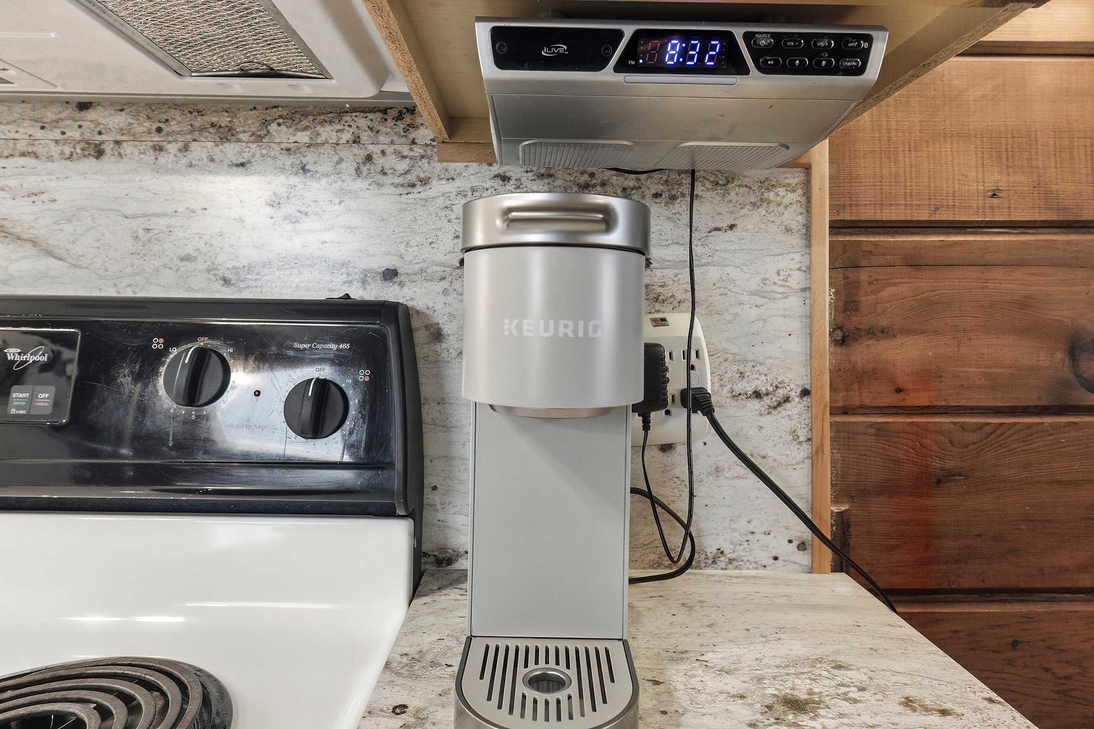 A close-up view of the kitchen counter highlights a modern grey Keurig coffee maker, a black electric stove, and a light granite-patterned countertop with a textured backsplash. An under-cabinet radio/clock is mounted above.