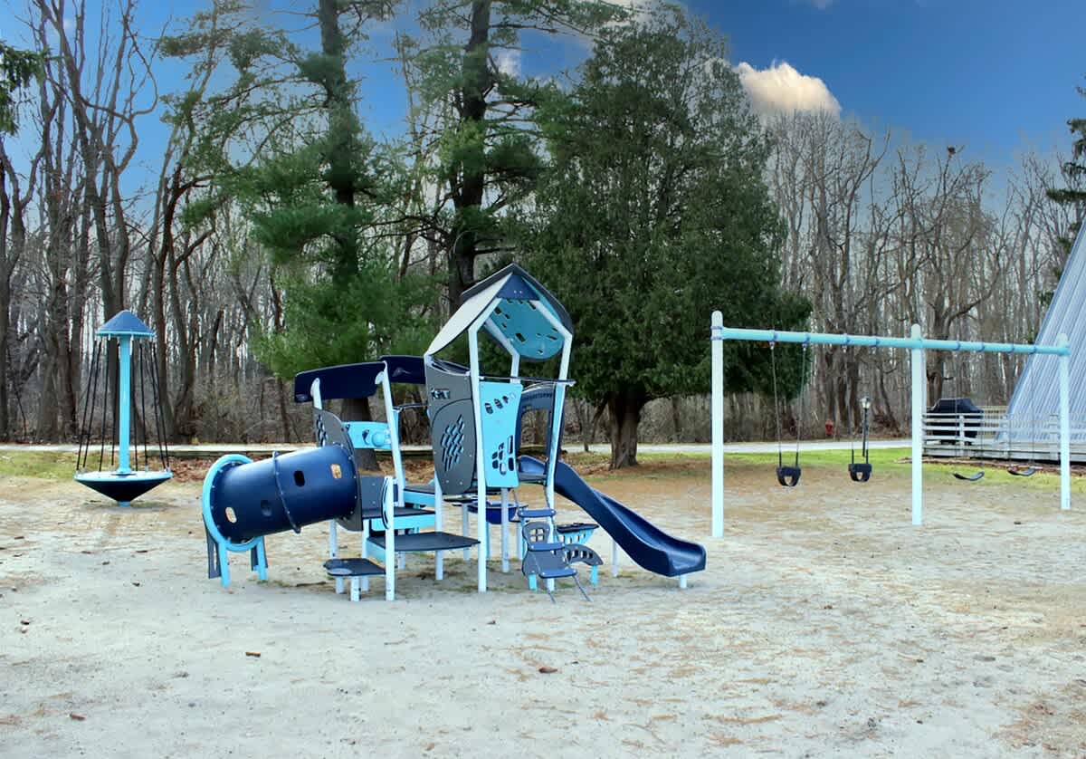 A modern blue and grey playground structure with slides and tunnels sits on a sandy base, flanked by swings and surrounded by tall trees, under a bright blue sky with white clouds.