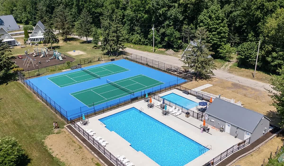Aerial view showcasing two outdoor swimming pools, one large and one smaller, adjacent to two blue and green tennis courts, all enclosed by fencing. A grey building is situated between the pools, surrounded by lush green grass and mature trees.