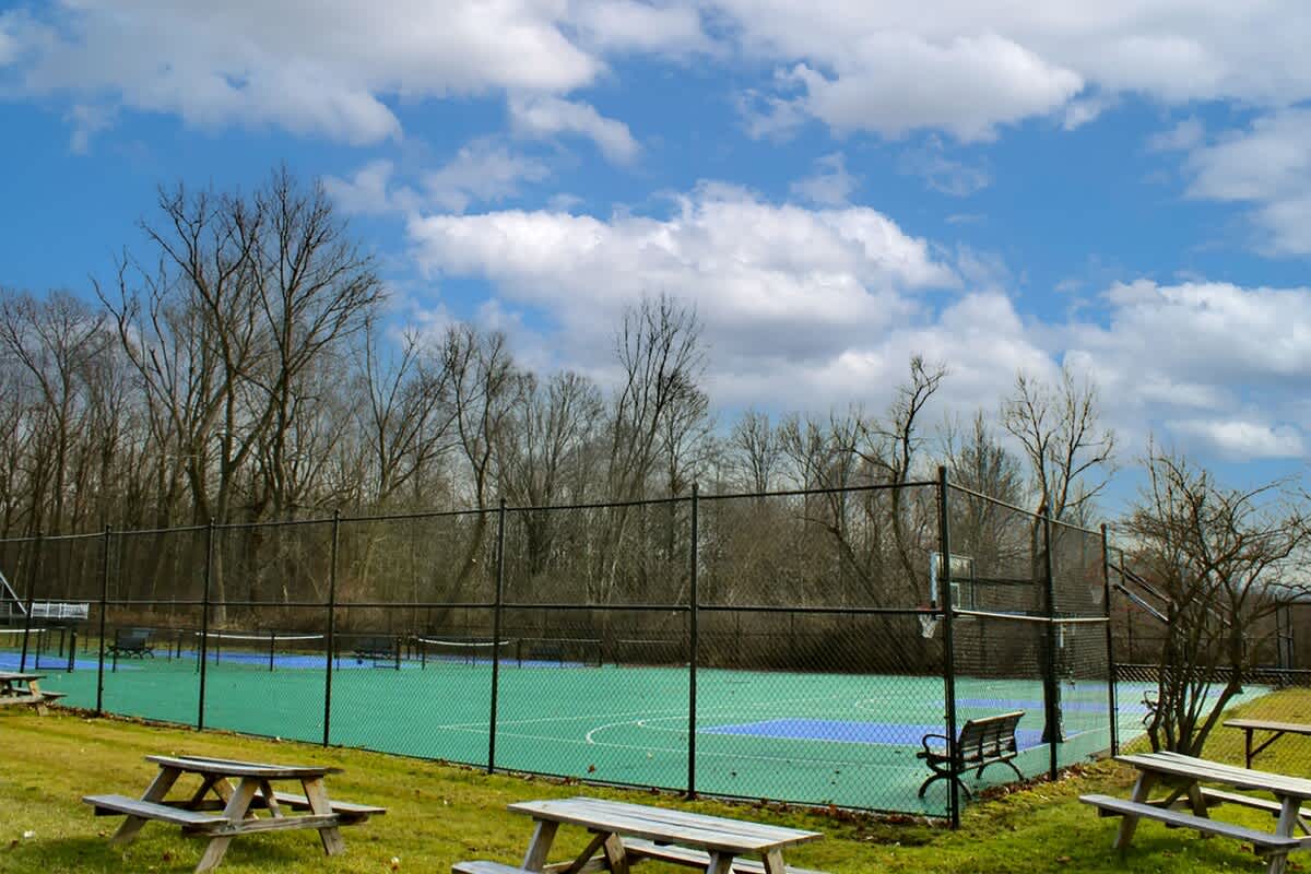 An outdoor sports court with a vibrant green and blue surface, enclosed by a chain-link fence, suitable for tennis or pickleball. Several wooden picnic tables are in the foreground, with a backdrop of bare trees under a partly cloudy sky.