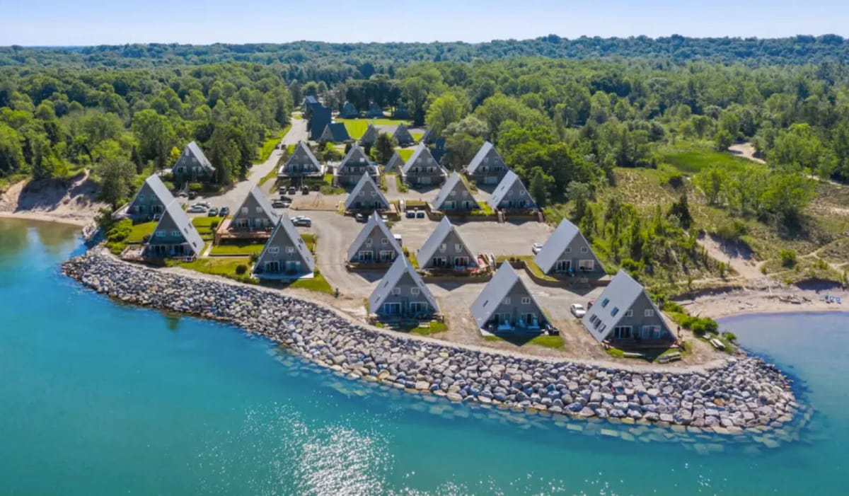 A wide aerial shot reveals a community of numerous A-frame houses arranged along a curved, rocky waterfront, with a sandy beach area and dense green forest extending into the background, all under a clear blue sky.
