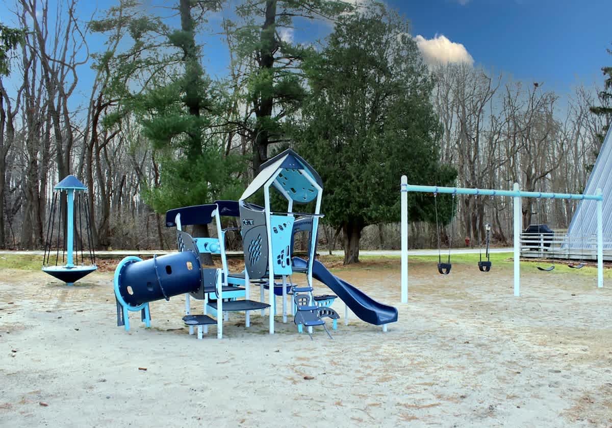 A well-equipped sandy playground features a blue and gray play structure with slides and tunnels, alongside a set of swings and a spinning toy, all nestled among mature trees.