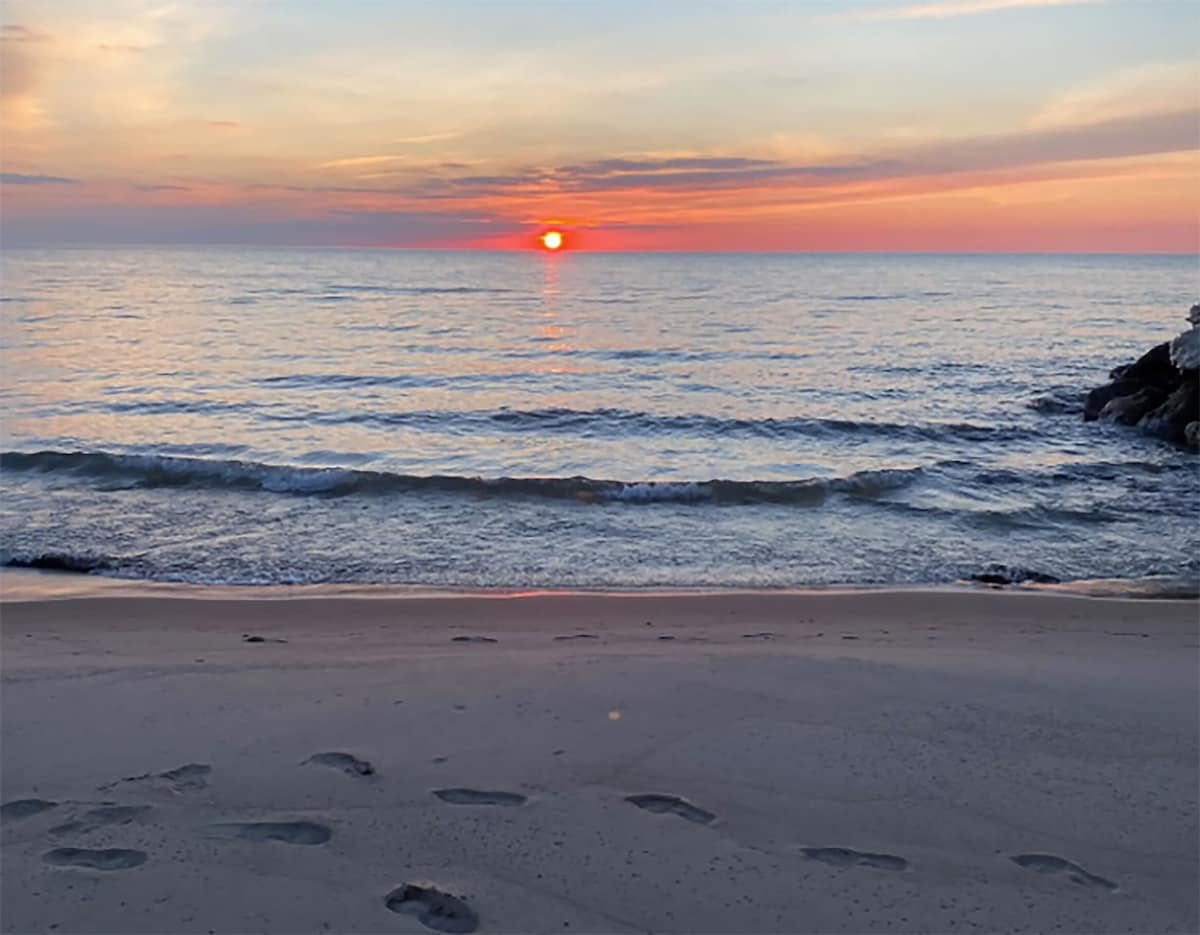 A serene sunset scene unfolds over a calm body of water, with the sun dipping below the horizon casting warm orange and red hues across the sky and water, viewed from a sandy beach with visible footprints.