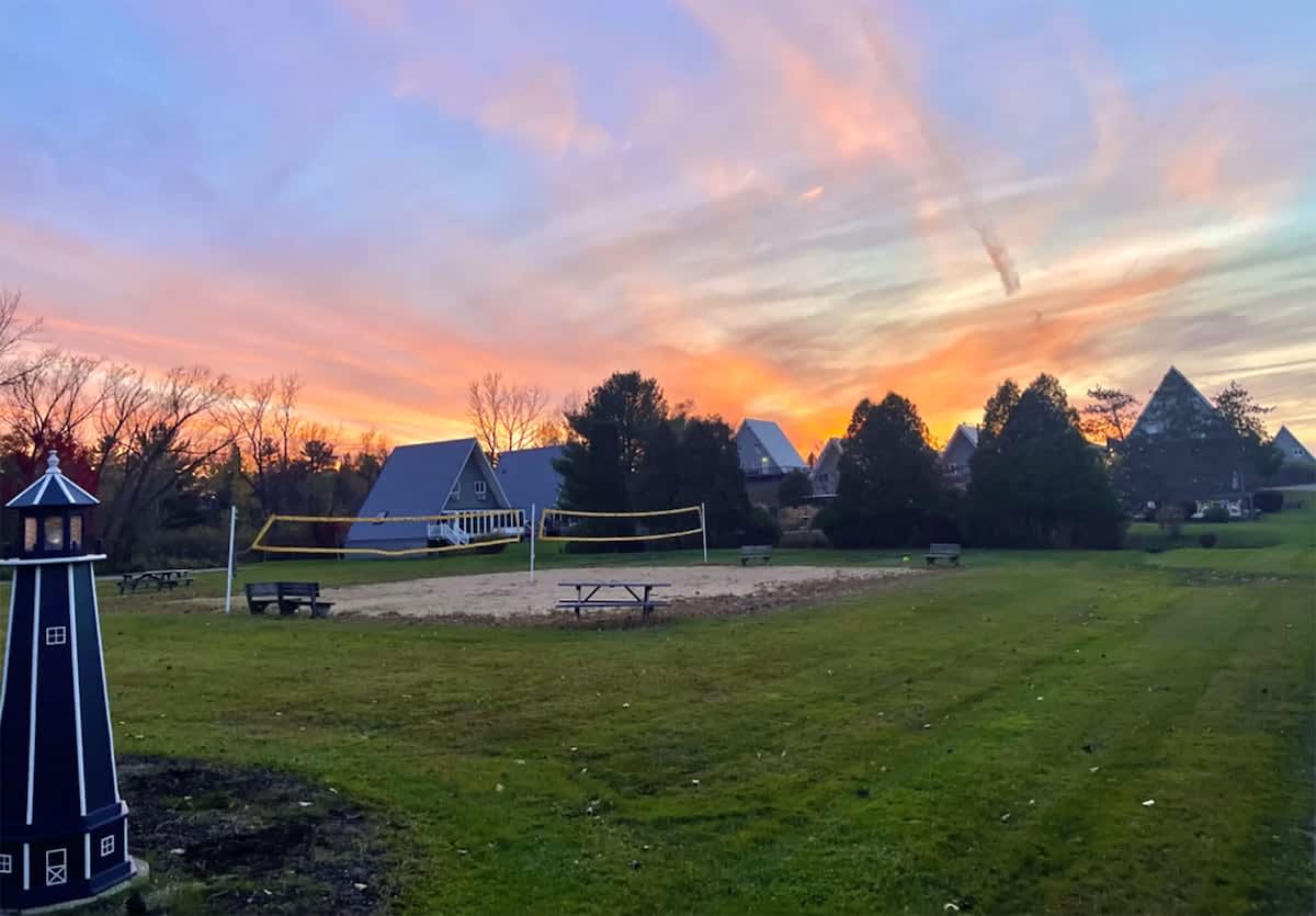 A sandy volleyball court with a net, flanked by wooden picnic tables and a charming decorative lighthouse, is set against a stunning sunset sky with vibrant orange and pink hues, with A-frame houses visible in the distance.