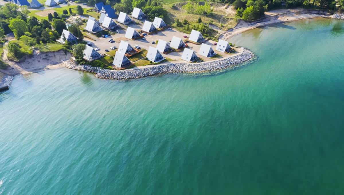 An aerial view showcases a cluster of distinctive A-frame houses nestled along a rocky shoreline, bordering a vibrant turquoise body of water, with lush green trees providing a natural backdrop.