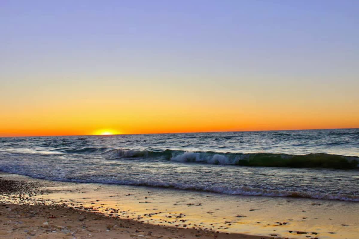 Vibrant sunset over a vast body of water, casting warm orange and yellow hues across the horizon, with gentle waves lapping onto a sandy and pebbled beach.