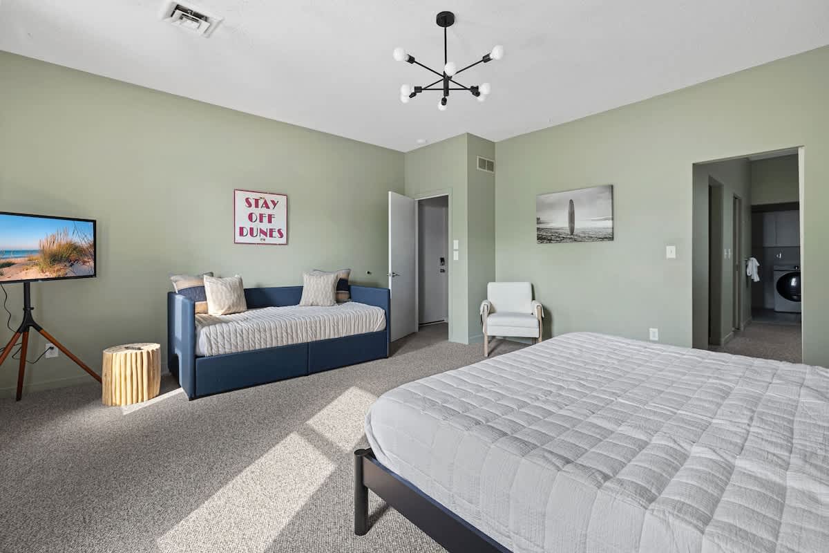 Spacious bedroom with light sage green walls, featuring a large bed with white bedding, a blue daybed with pillows, a wall-mounted TV on a tripod stand, and a modern black chandelier.
