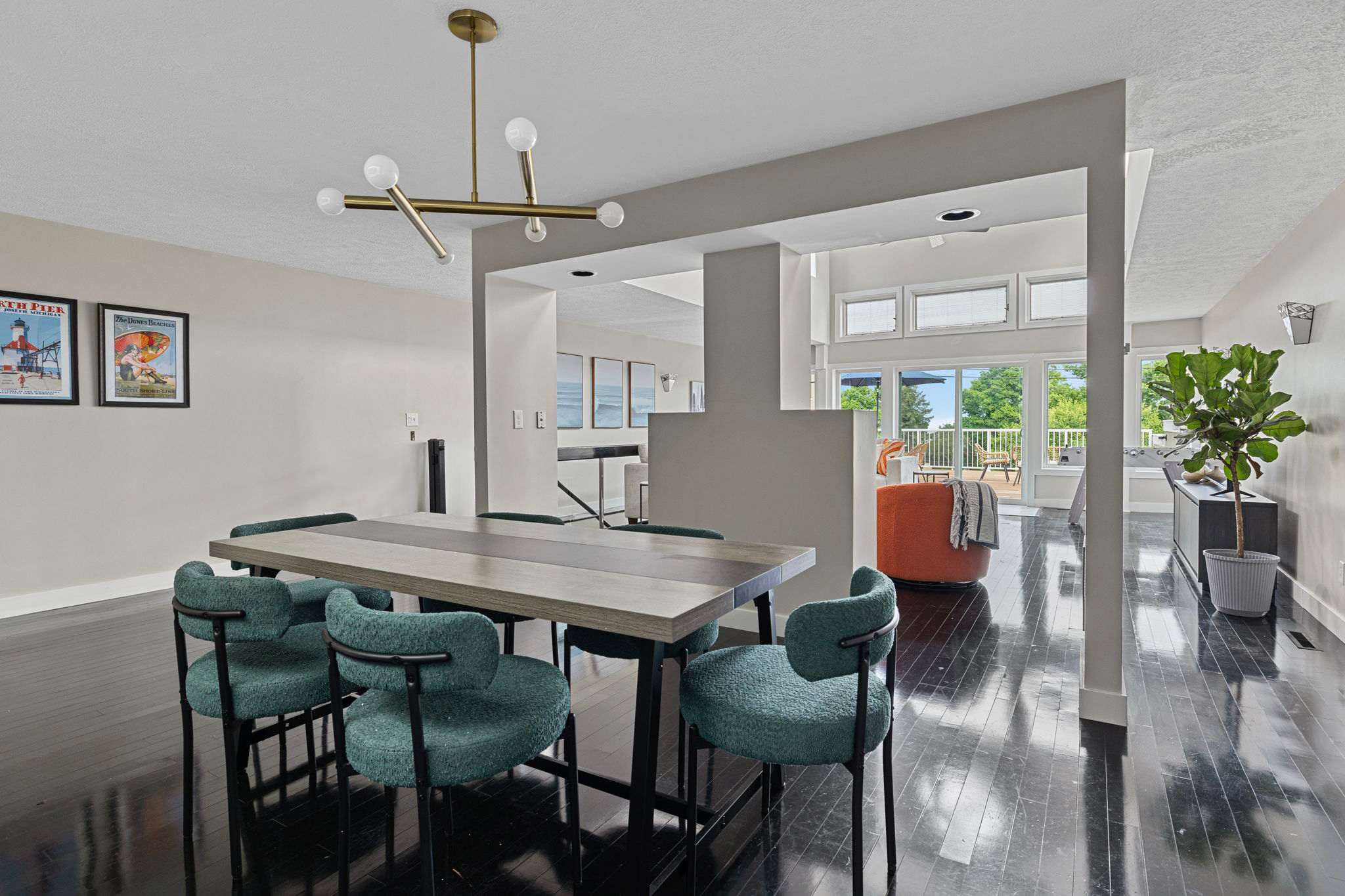 Modern dining area featuring a long rectangular table with six teal upholstered chairs on dark hardwood floors, illuminated by a linear brass chandelier. The space opens to a bright living area and an outdoor deck.
