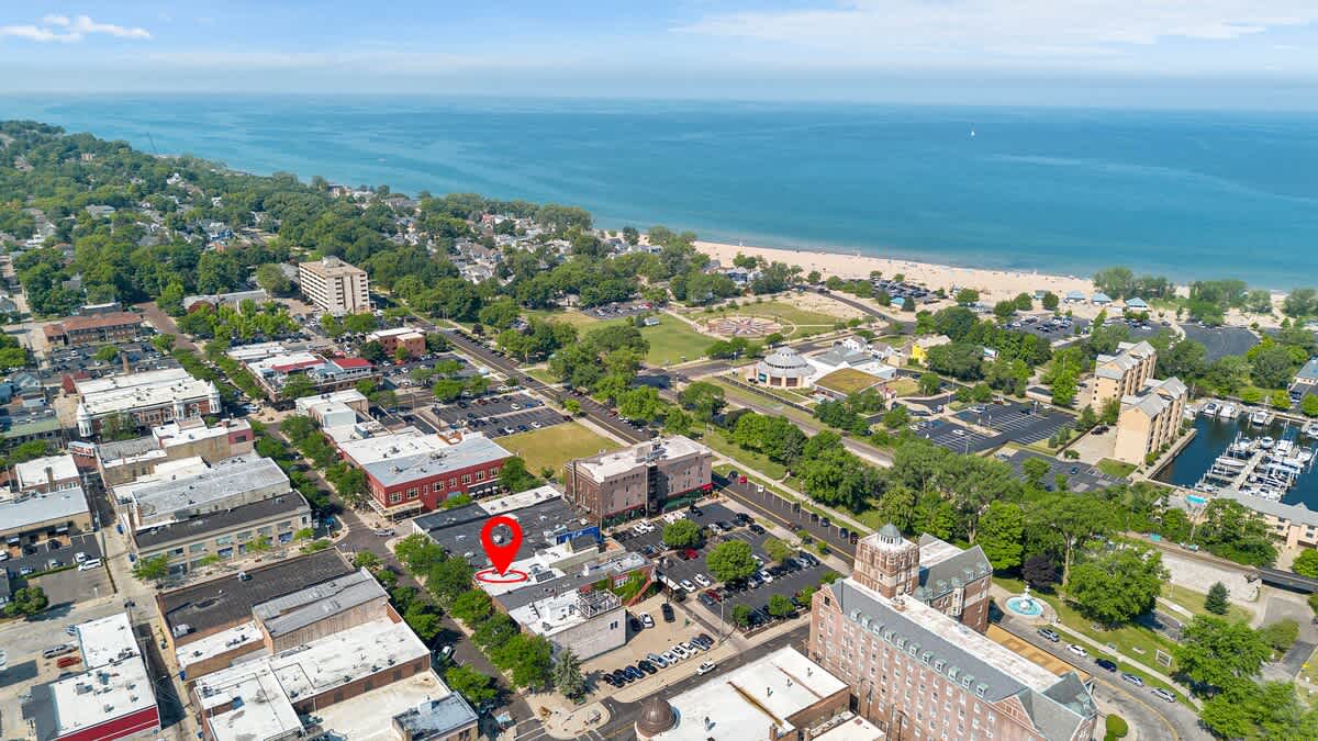 An expansive aerial view of a vibrant coastal town, showcasing the property marked with a red pin amidst commercial buildings, with a wide sandy beach and the vast blue lake stretching into the horizon under a bright sky.