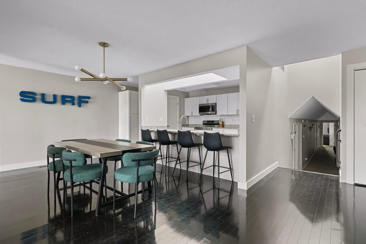 Open-concept dining area with a long table and teal chairs, adjacent to a kitchen bar with black stools, all set on dark hardwood floors. A large 'SURF' sign decorates the wall, leading into a hallway.