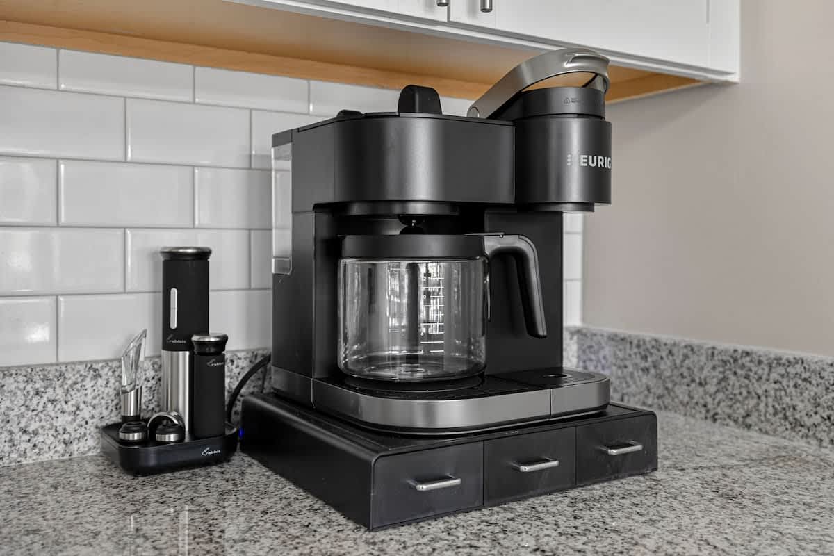 Close-up of a black Keurig coffee maker and a wine opener set on a light granite countertop, against a white subway tile backsplash, showcasing kitchen amenities.