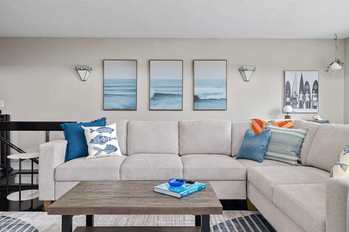 Detailed view of the light gray sectional sofa adorned with blue and patterned throw pillows. Three framed ocean wave artworks hang above the sofa, complemented by wall sconces and a dark wood coffee table.