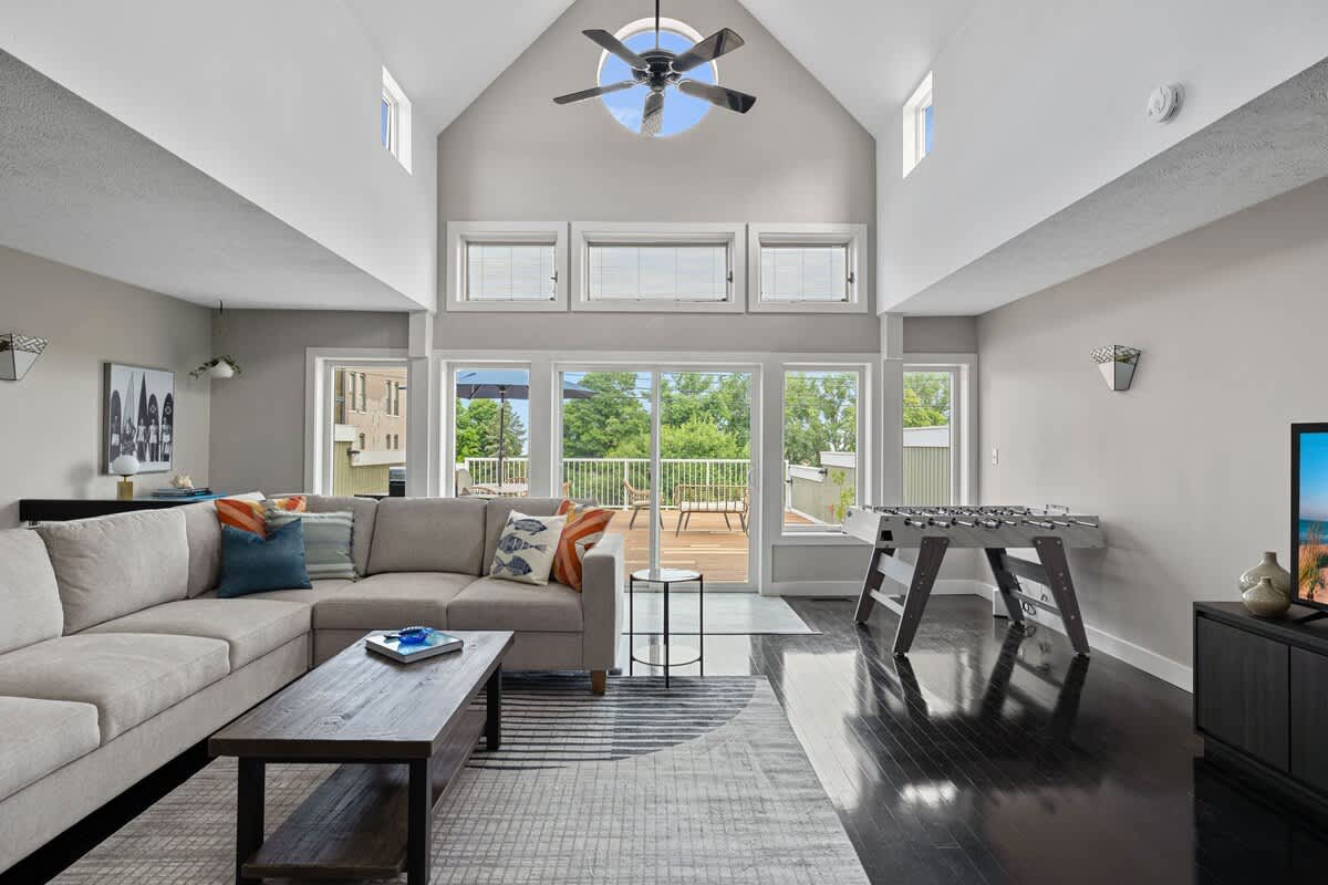 Expansive living room with a light gray sectional sofa, dark wood coffee table, and a foosball table. High vaulted ceilings with a ceiling fan and skylights illuminate the space, which opens to an outdoor deck through large glass doors.