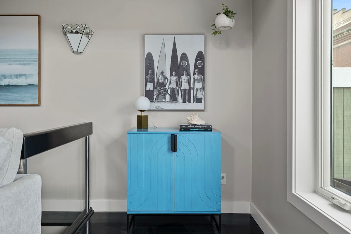 A vibrant blue cabinet with arched details stands against a light gray wall, topped with a white lamp and a decorative shell, beneath a framed black and white surf-themed print. A small hanging plant adds greenery to the corner.