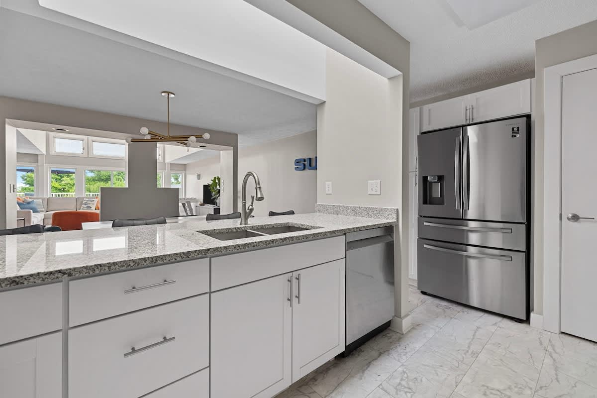 Modern kitchen with white cabinets, light granite countertops, and stainless steel appliances including a large refrigerator and dishwasher. A sink with a gooseneck faucet is integrated into the counter, with light tile flooring.