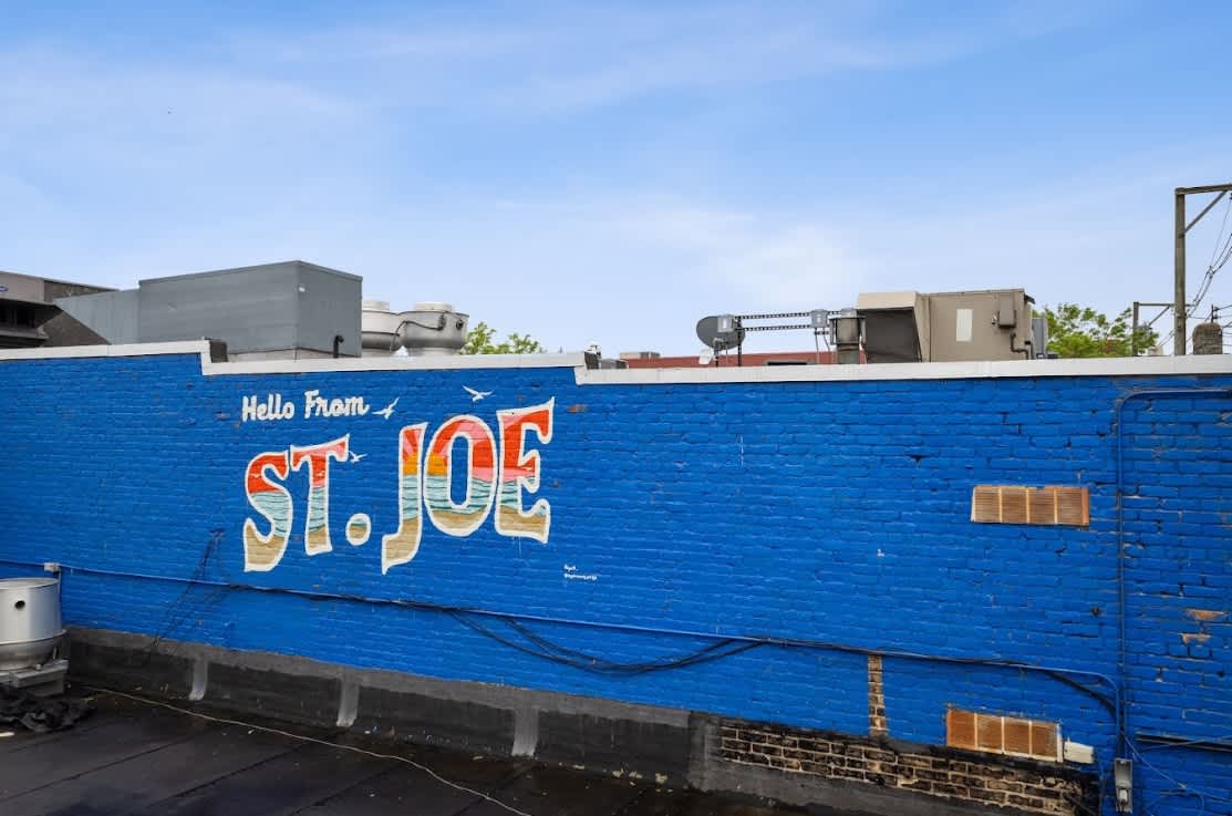 Rooftop view featuring a vibrant blue brick wall with a 'Hello From St. Joe' mural, surrounded by various rooftop vents and equipment under a clear sky.