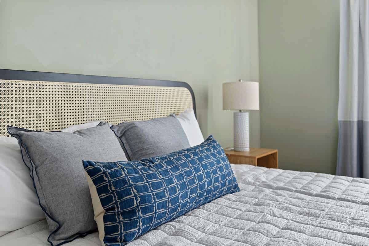 Close-up of a bed featuring a stylish cane and black frame headboard, adorned with blue and grey decorative pillows, and a white quilted comforter, beside a wooden nightstand with a lamp.