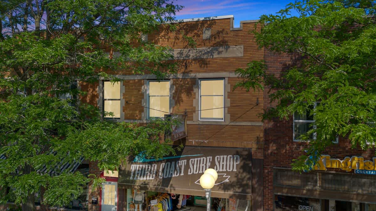 Street-level exterior view of the brick building, featuring large windows and a "Third Coast Surf Shop" sign on the ground floor. Lush green trees partially obscure the facade.
