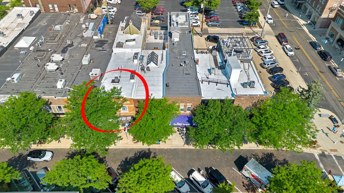 A detailed aerial perspective of the property, highlighted by a red circle, revealing its rooftop and the surrounding urban landscape of commercial buildings, bustling streets with parked cars, and lush green trees.