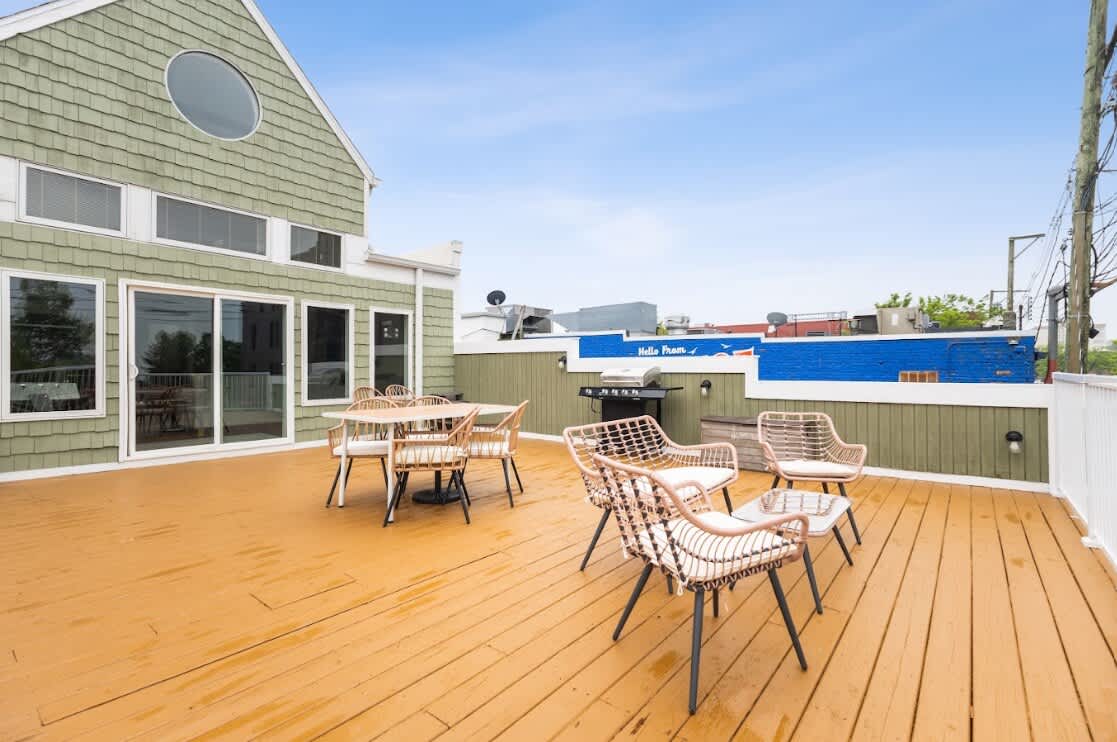 Sunny rooftop deck with a wooden floor, featuring an outdoor dining set with an umbrella and a separate lounge area with wicker chairs. The building's green shingled exterior and a white railing are visible.