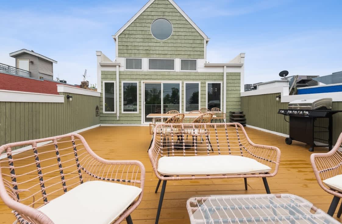 Spacious outdoor deck with light pink woven patio furniture and a dining set, offering views of a green-sided house with large windows and a round attic window. A BBQ grill is visible on the right, ready for outdoor cooking.