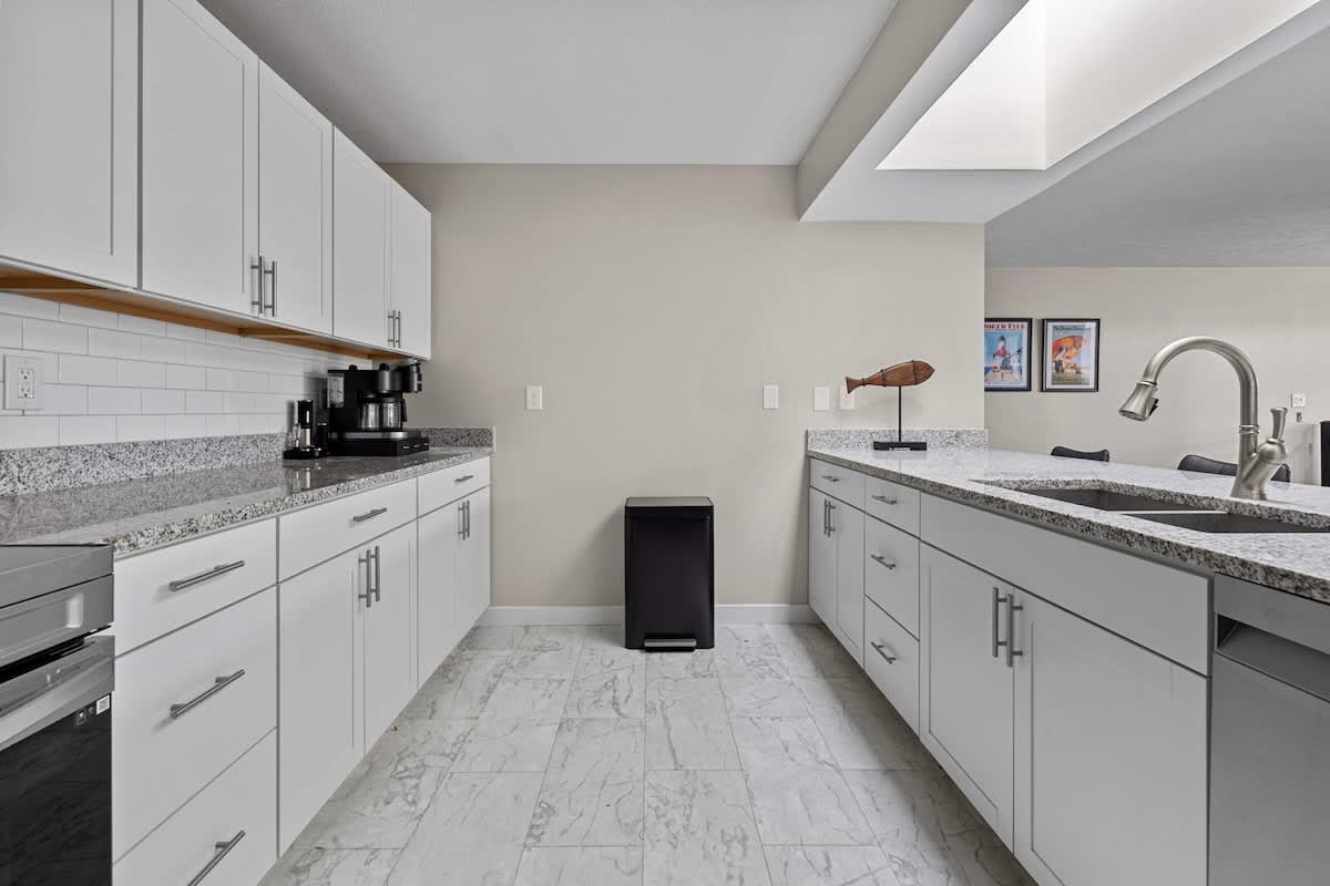 Galley-style kitchen with white shaker cabinets, light granite countertops, and a white subway tile backsplash, featuring a coffee maker and a black trash can. The floor is covered in light-colored marble-look tile.