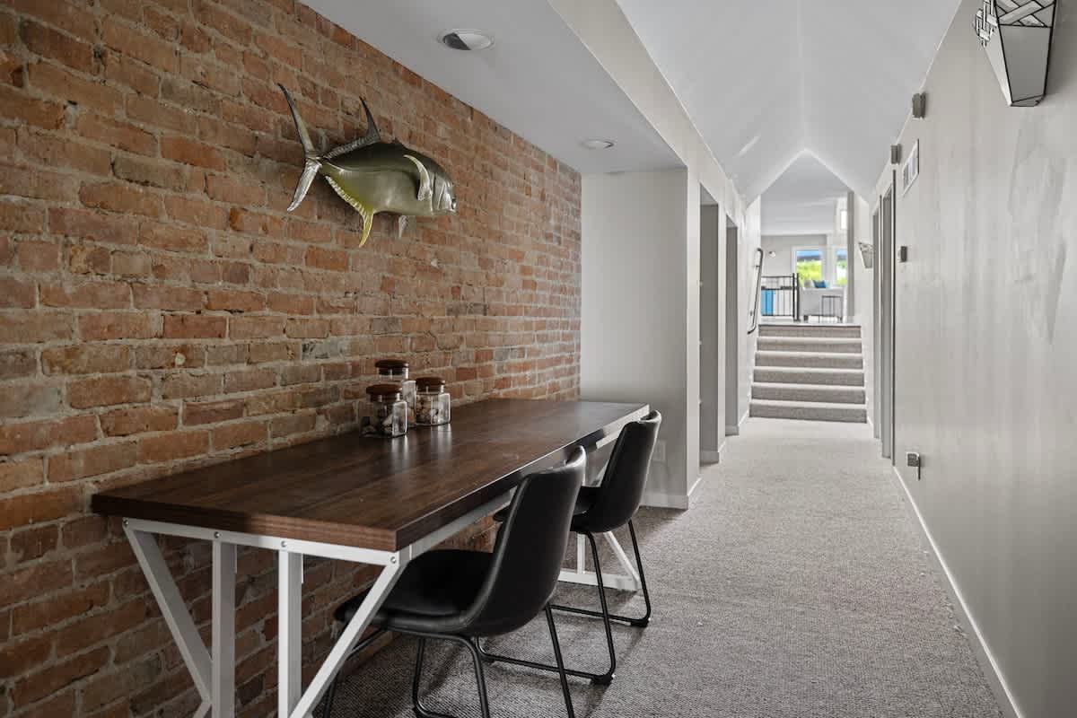 Industrial-style hallway or workspace featuring an exposed brick wall with a mounted fish decoration. A long wooden table with two black chairs provides a functional area, with carpeted stairs visible in the distance.