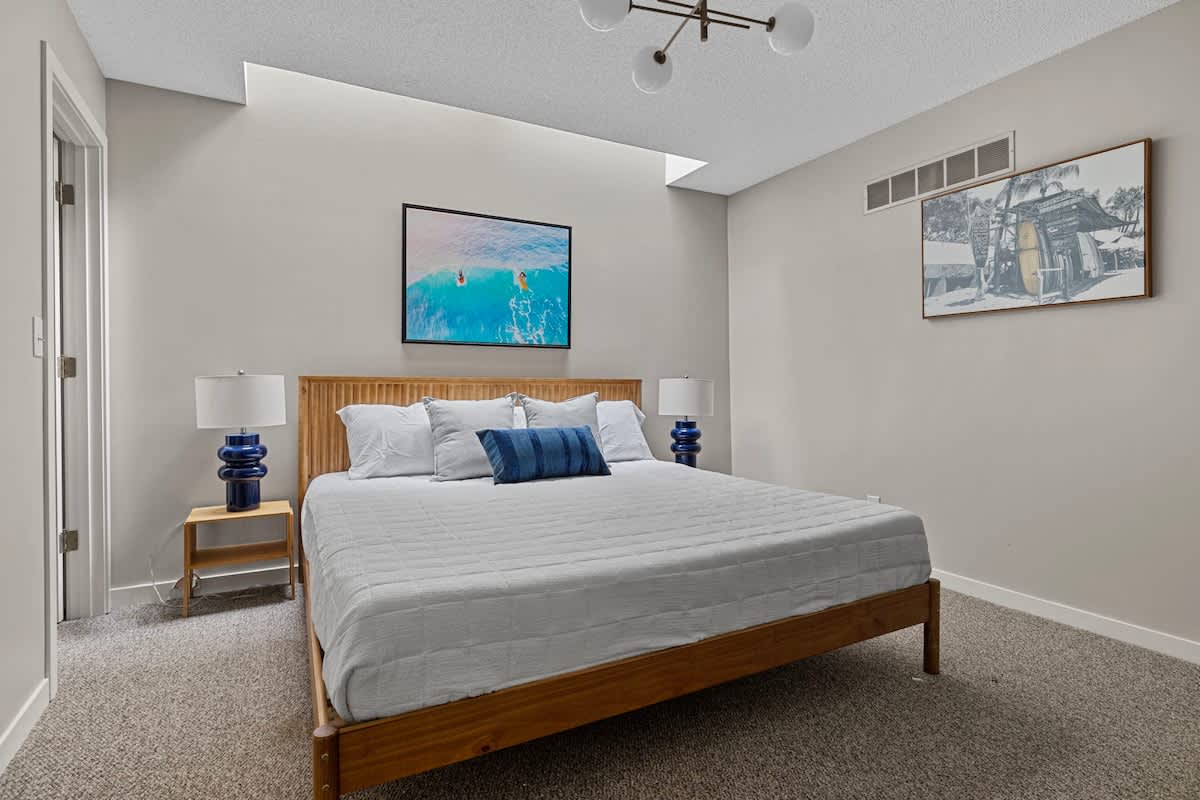 Bright bedroom with light grey walls, featuring a wooden platform bed with white bedding, two blue ceramic lamps on wooden nightstands, and modern artwork above the bed.