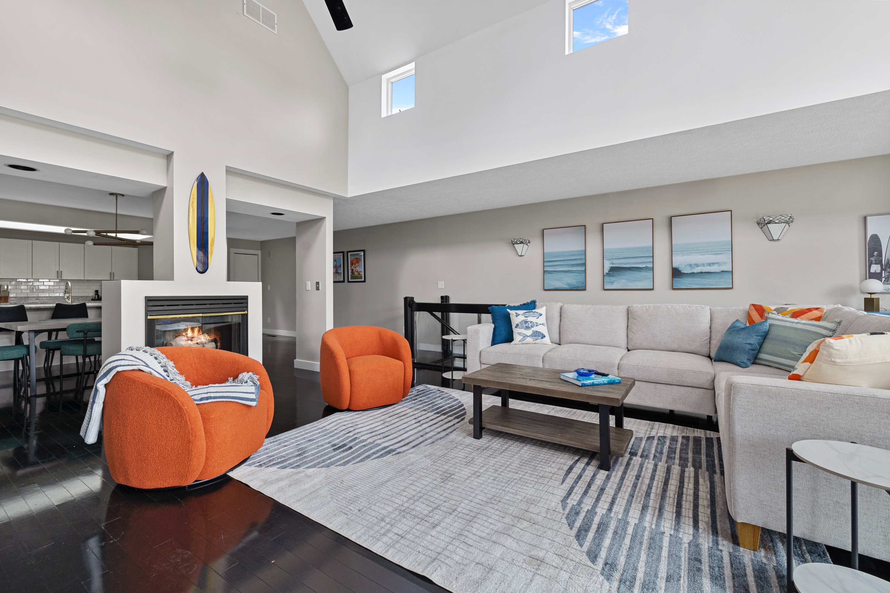Cozy living room with a modern fireplace flanked by two vibrant orange armchairs. A large light gray sectional sofa and a dark wood coffee table sit on a striped area rug, under high vaulted ceilings with skylights.