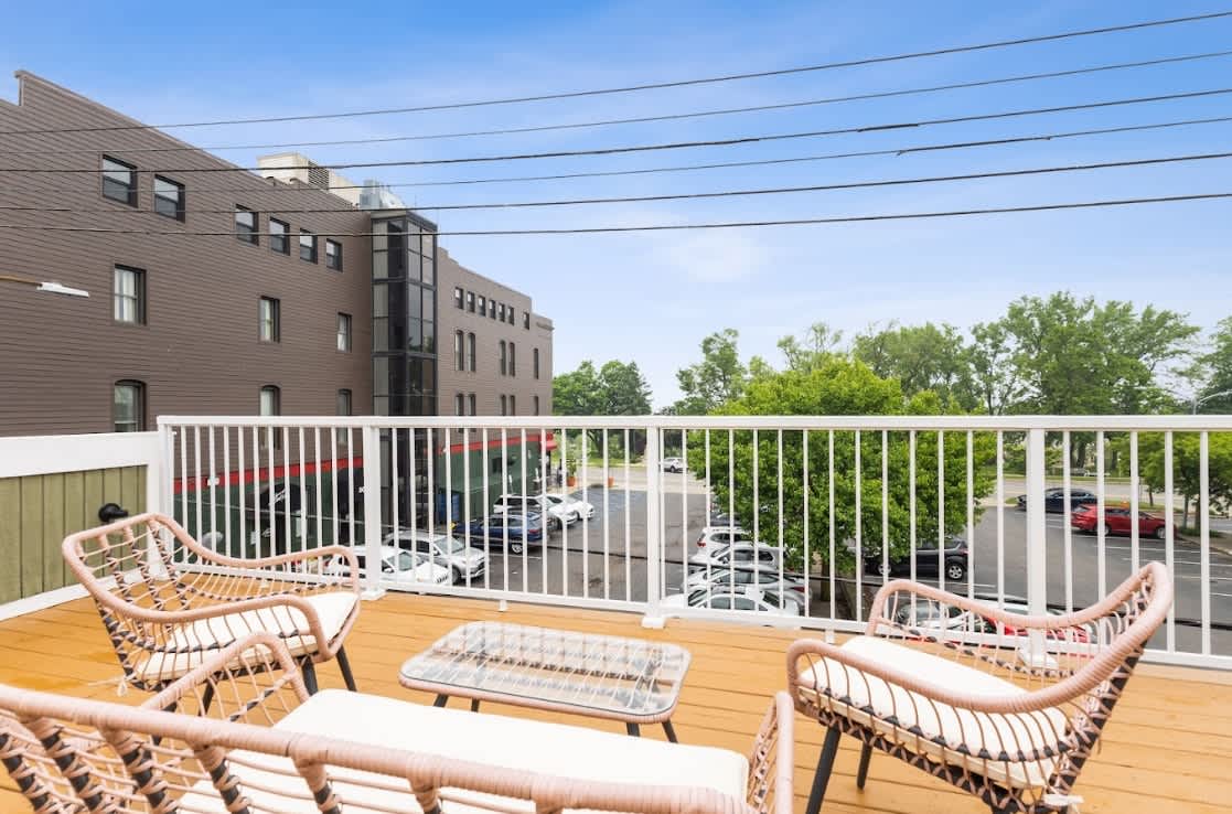 Outdoor balcony with a warm wooden deck floor, white railing, and a casual seating area including two wicker chairs and a glass-top coffee table, overlooking a parking lot and adjacent buildings.