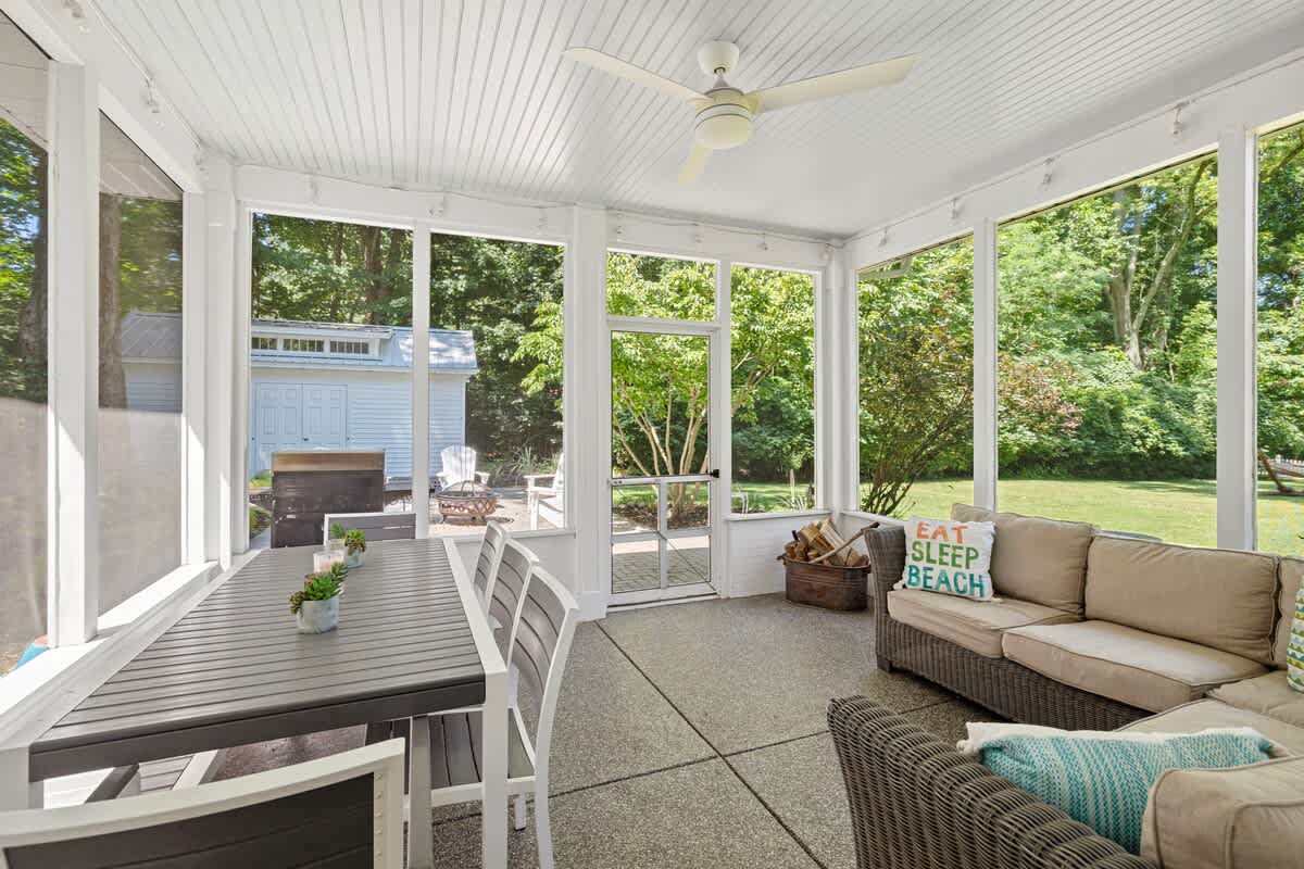 A bright screened porch features a large dark gray dining table with white chairs on one side and a wicker sectional sofa with light cushions on the other, all under a white paneled ceiling with a fan. Large screens offer views of the surrounding greenery.