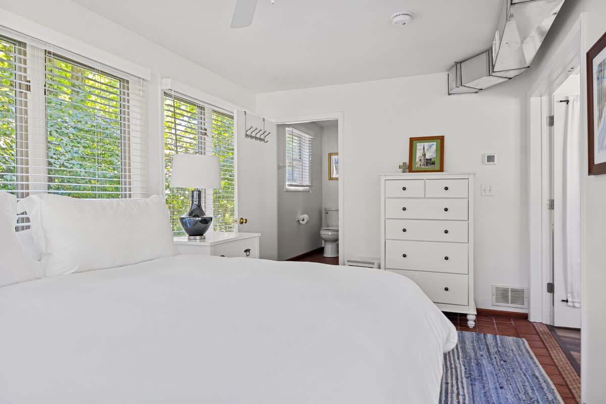 View of a bedroom with a white bed, white dresser, and terracotta tile flooring. A window with blinds provides natural light, and an open doorway leads to an en-suite bathroom.