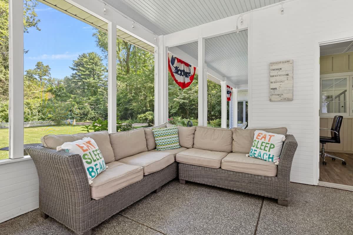 A spacious screened-in porch featuring a large wicker sectional sofa with light cushions and 'EAT SLEEP BEACH' pillows. The porch overlooks a lush green yard and trees, with a patriotic banner visible in the background.
