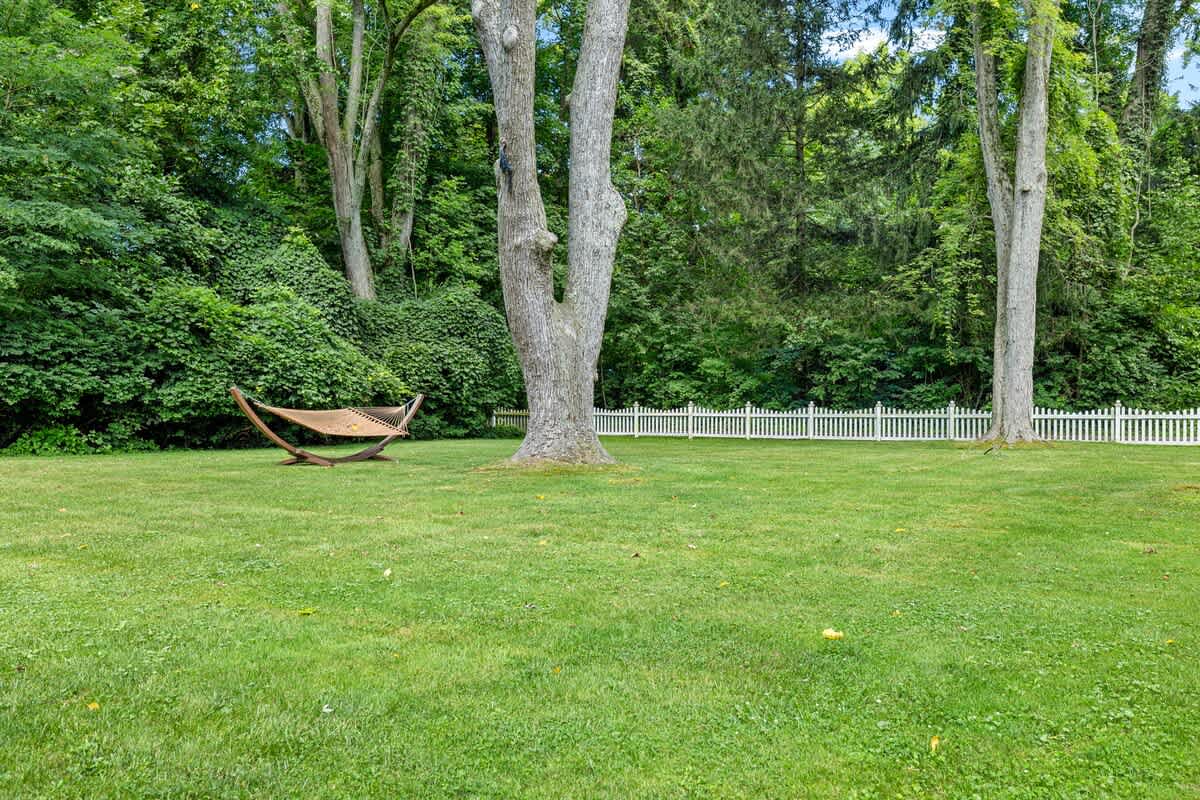 A large, well-maintained green lawn with several mature trees providing ample shade. A wooden hammock with a stand is set up in the middle of the yard, and a white picket fence lines the back.