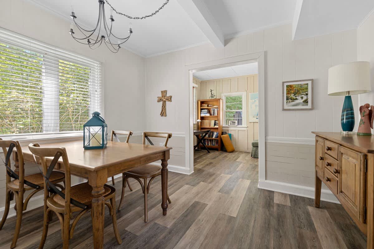 Bright dining room with light wood plank flooring, a rectangular wooden dining table with four chairs, and a large window with blinds. A dark metal chandelier hangs from the ceiling, and a wooden cross decorates one wall.