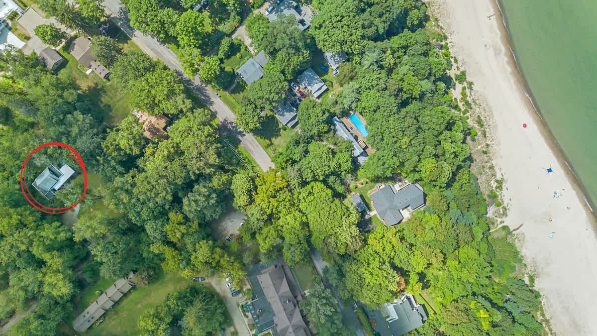 An aerial view showing the property, circled in red, nestled among green trees, with a road and other houses visible, and a sandy beach bordering a large body of water in the upper right.