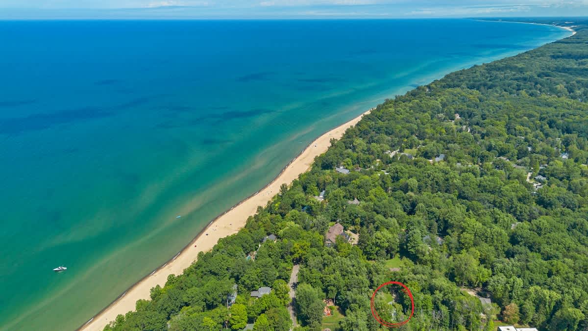 A panoramic aerial view highlighting the property's location, circled in red, within a dense green forest, with the vibrant blue waters of a large lake or ocean and a wide sandy beach in the foreground.