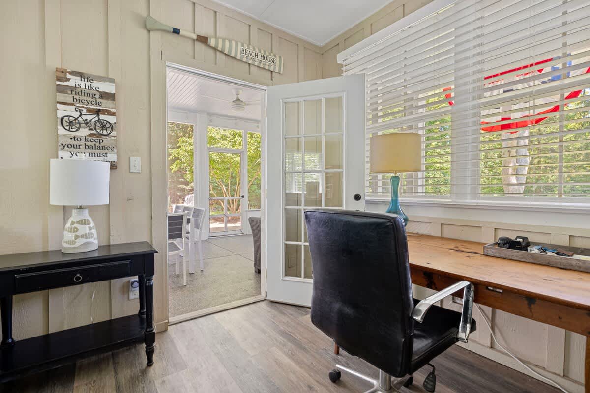 Dedicated office space with a wooden desk, black office chair, and light wood plank flooring. A large window with blinds overlooks greenery, and a glass-paned door leads to a screened porch.