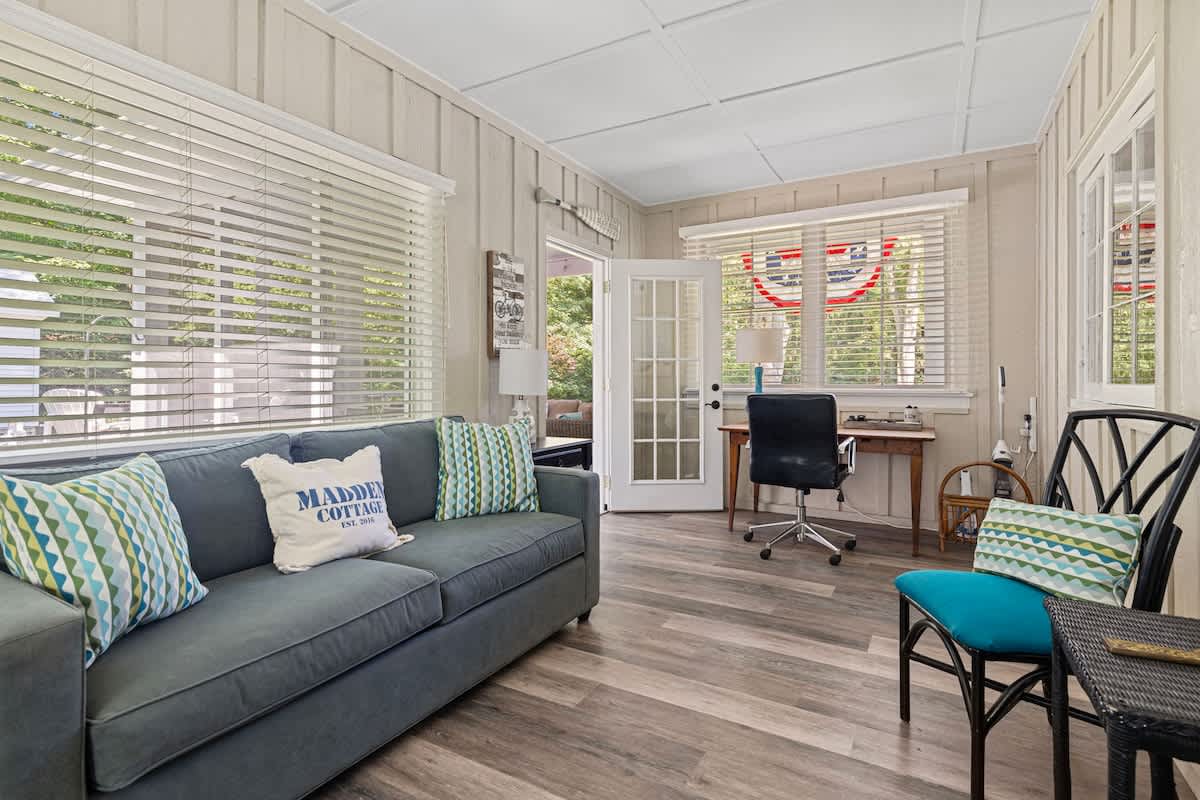A cozy sunroom features a dark blue sofa with patterned pillows, light wood-paneled walls, and large windows with white blinds. A small wooden desk with an office chair is tucked into a corner, offering a dedicated workspace.