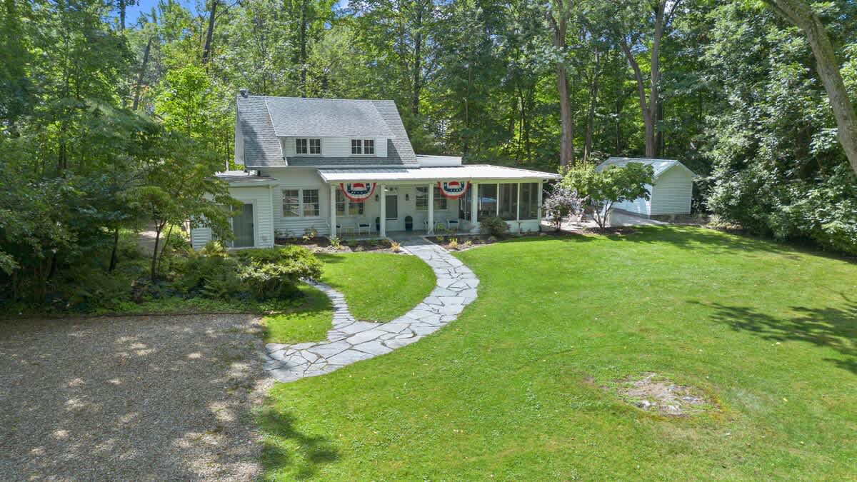 A wider exterior view of the white clapboard house with a gray roof, a covered porch decorated with patriotic bunting, a stone pathway, and a spacious green lawn, all surrounded by dense, mature trees.