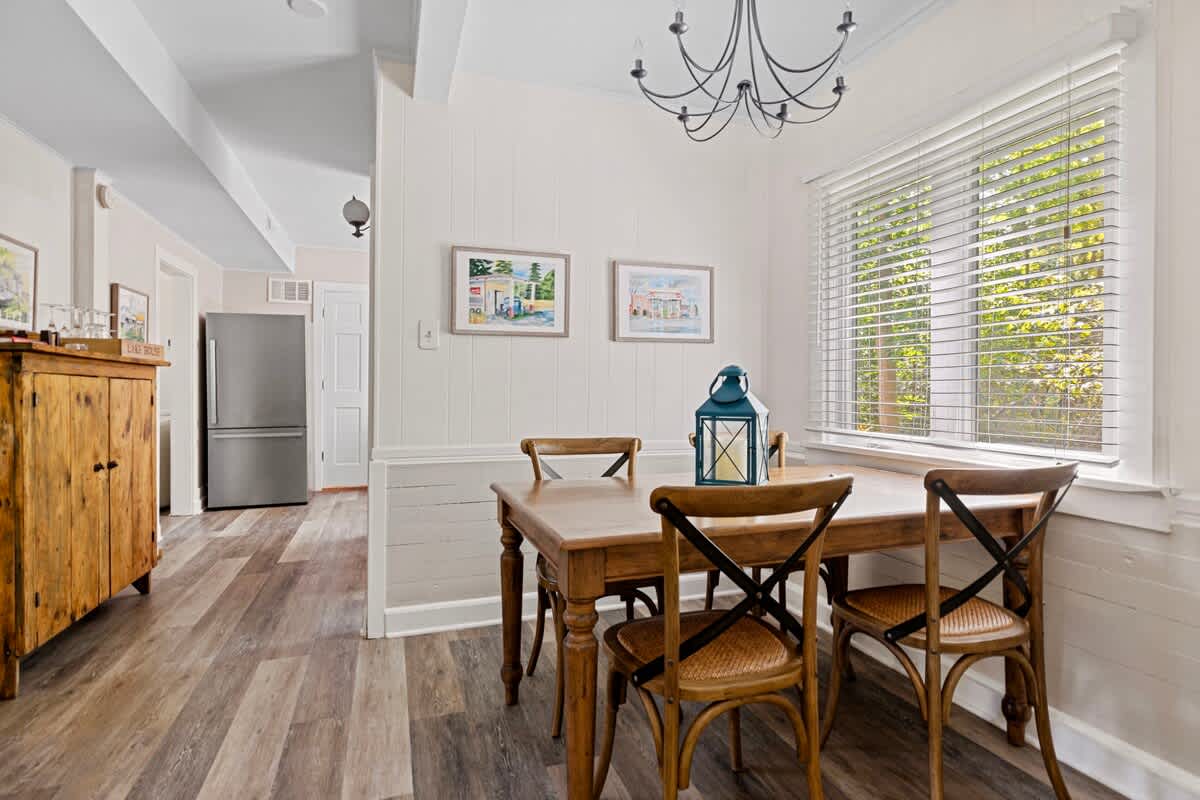View of the dining area with a wooden table and chairs, light wood plank flooring, and a large window. A wooden cabinet with drawers and a lamp sits against the wall, and an open doorway leads to another room.
