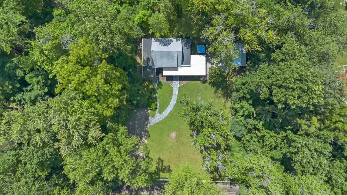 An overhead aerial view of the white house with a gray roof, featuring a stone pathway and a small shed in the backyard, completely enveloped by a lush, green tree canopy under a bright sky.