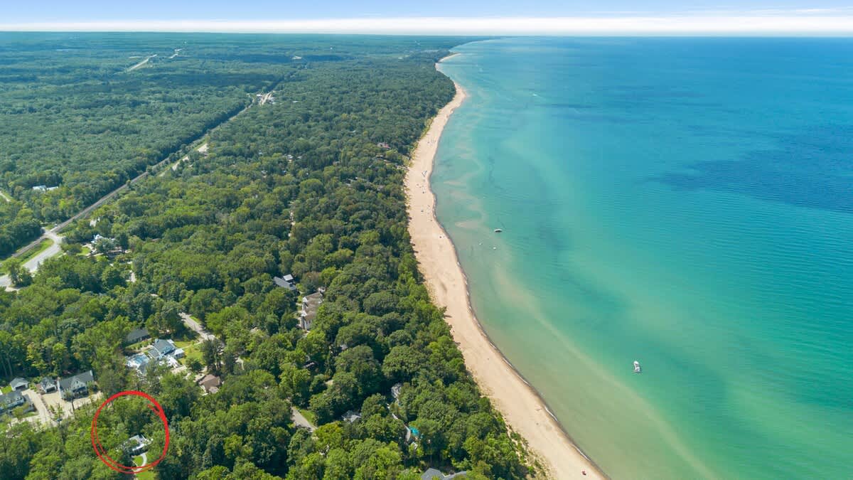 An expansive aerial view showcasing the property, circled in red, surrounded by dense green forests, with a vast blue body of water and a long sandy beach stretching along the coastline in the background.