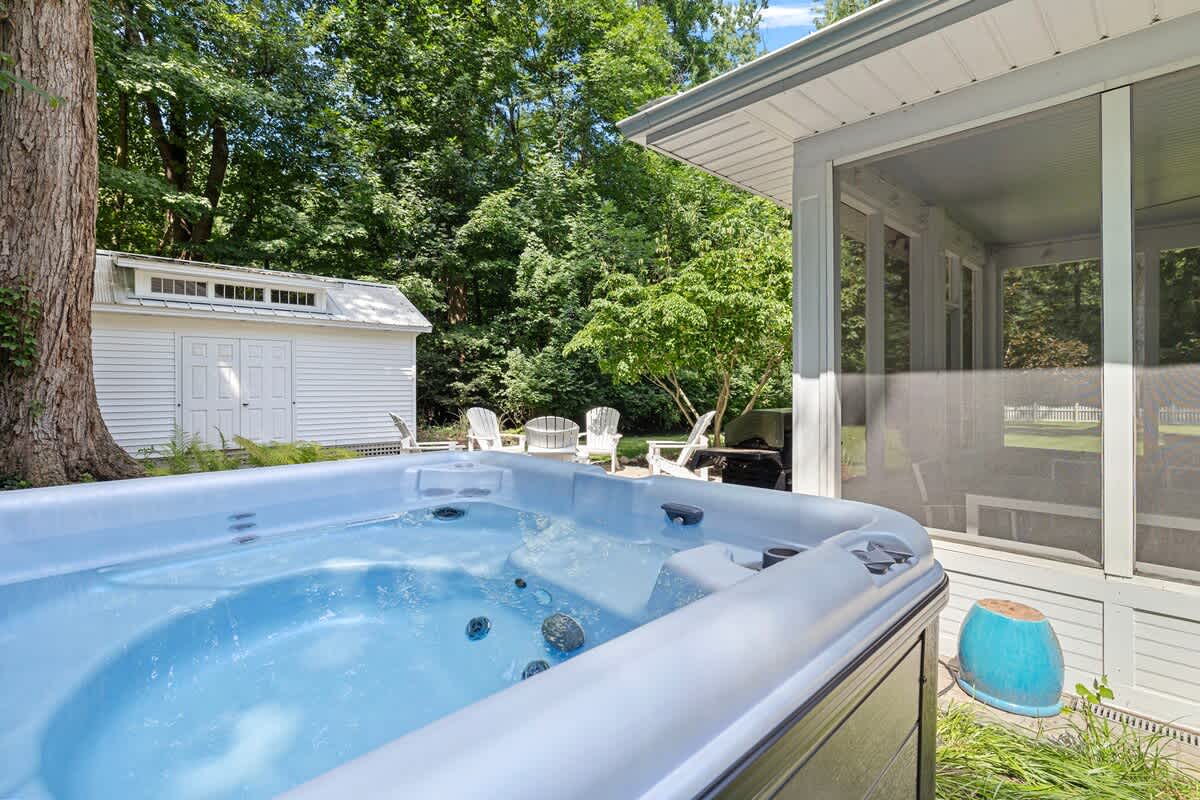 A close-up view of a grey hot tub filled with sparkling blue water, situated in a green yard. In the background, a white shed, white Adirondack chairs, and a white picket fence are visible among tall trees.