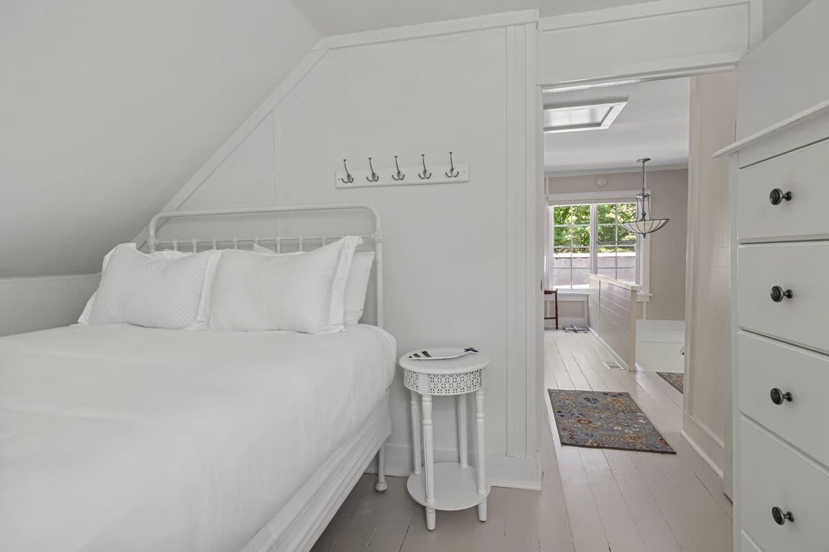 A cozy bedroom with white paneled walls and a white metal-frame bed with white bedding. A small white round nightstand sits beside the bed, and a row of hooks is mounted on the wall.