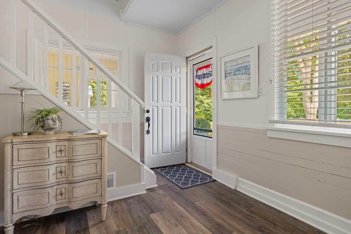 The bright foyer features light wood-look flooring, white wainscoting, and a white front door with a screen door. A small white chest with a plant on top sits beside a staircase, with a blue patterned rug at the entrance.