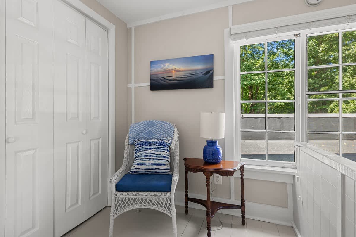 Bright corner with a white wicker chair, blue cushion, and a small wooden side table with a lamp. A large window offers views of trees, and a white bi-fold closet door is visible.