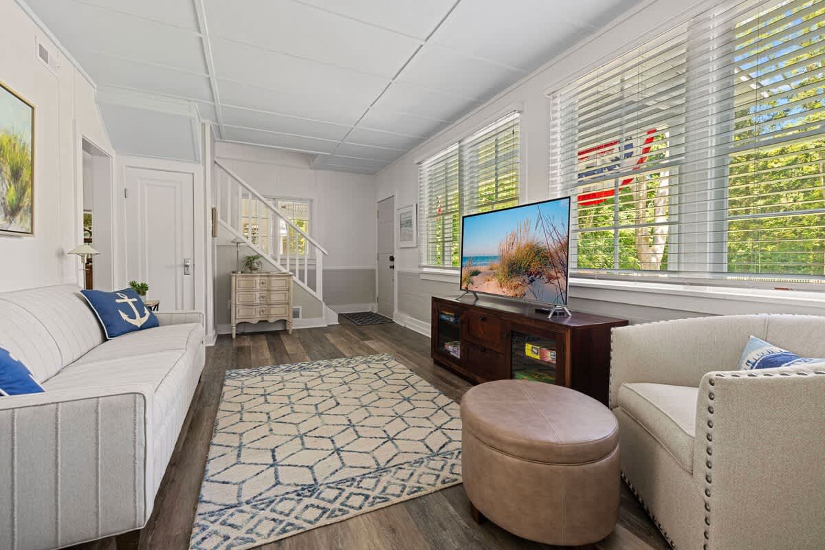 The living room features light wood-look flooring and white paneled walls, with a light gray sofa and two armchairs arranged around a patterned area rug. A large flat-screen TV sits on a dark wooden console, positioned beneath windows with white blinds.