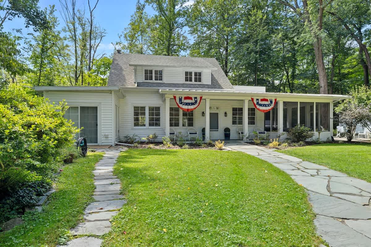 The front exterior of a charming white clapboard house with a gray roof, featuring a covered porch adorned with patriotic red, white, and blue bunting, and a winding stone pathway leading through a lush green lawn.