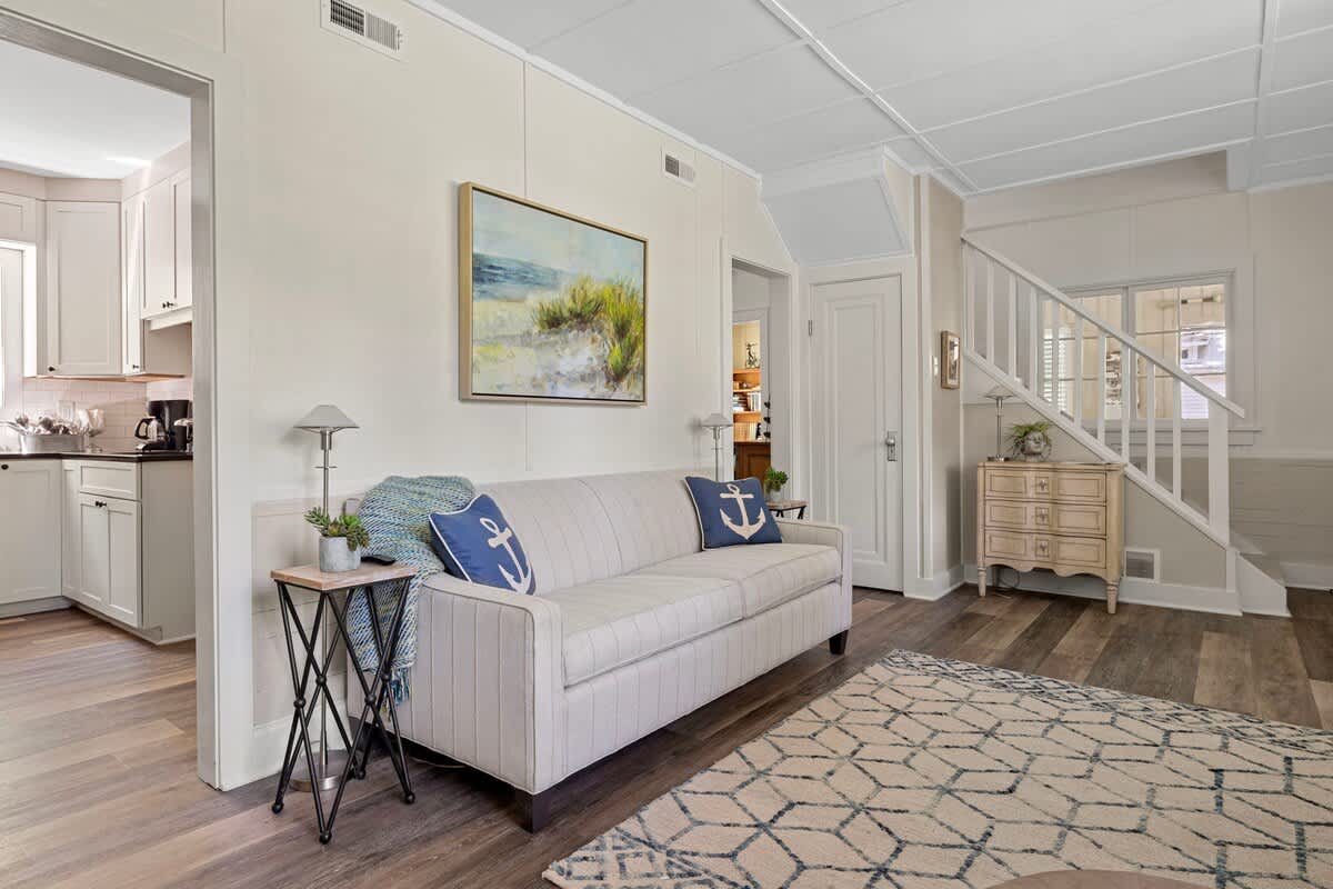 A light and airy living room features wood-look flooring, white paneled walls, and a light gray sofa with blue anchor pillows. A small side table with a lamp is next to the sofa, and an open doorway leads into a modern kitchen.